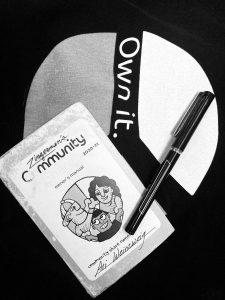 black and white photo of a Zingerman's Community Shares owner's manual, a ballpoint pen, and a t-shirt that says "Own It"